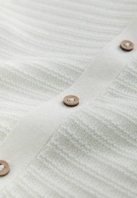 Knapp-front stickad tröja i krämfärg; har ett texturerat mönster med vertikala ränder och träknappar längs mitten.