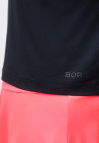 Κοντινό πλάνο στο τελείωμα ενός μαύρου πουκαμίσου με το λογότυπο "BORG" πάνω από φούστα ή σορτς φωτεινού ροζ χρώματος.