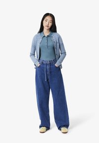 Ljusblå kabelstickad cardigan över en teal polo, tillsammans med vida mörkblå denimjeans och ljusa sneakers.