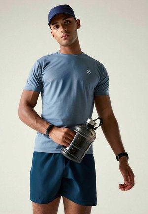 Athletic man wearing blue cap, t-shirt, and shorts holds large water bottle, standing against a plain light background.