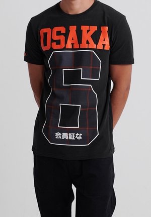 Czarny bawełniany t-shirt z wyrazistym czerwonym napisem „OSAKA” i dużą kraciastą cyfrą „6” na przodzie oraz białym japońskim tekstem poniżej.