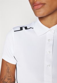 Polo bianco con un design aderente, dotato di colletto classico, bottoni trasparenti e un accento nero sulla spalla.
