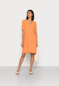 Vestido laranja de mangas curtas com decote em V, adornado com bolinhas brancas. Tecido leve, até ao joelho, combinado com uma mala bege e sandálias.