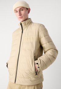 Jachetă puffer bej cu guler înalt, textură matlasată și buzunare cu fermoar. Are un logo subtil embosat pe față.