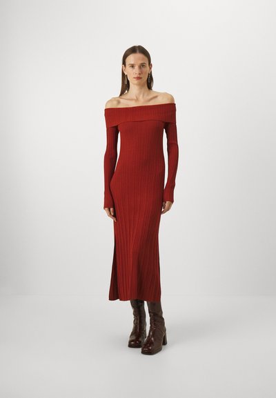 Rochie roșie cu nervuri, fără umeri, cu mâneci lungi, având o siluetă ajustată și un material texturat. Poartă cizme cu toc maronii.