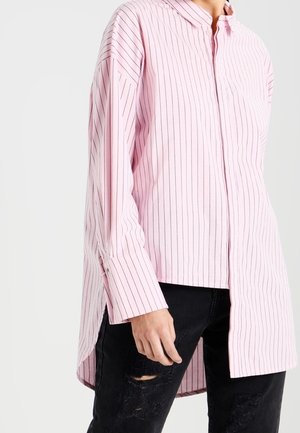 Personne portant une chemise rose à manches longues avec des rayures verticales et un ourlet asymétrique, ainsi qu'un jean noir déchiré, debout devant un fond uni.
