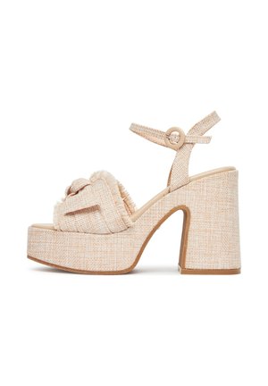 Sandalias de plataforma beige con parte superior de tela texturizada, detalle anudado y tacón grueso tipo bloque. La correa del tobillo presenta un adorno con hebilla redonda.