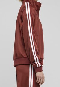 Maroon zip-up tracksuit jacket with white stripes along the sleeves, made of smooth fabric, featuring a stand collar and side pockets.