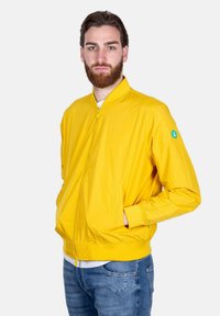 Giacca bomber gialla con zip frontale, polsini e vita in maglia, dotata di tasche laterali e una toppa con logo verde sulla manica sinistra.