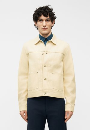 Mann mit lockigem Haar trägt eine cremefarbene Jacke mit Knöpfen über einem dunkelblauen Hemd mit Kragen und marineblaue Hose, steht vor einem einfarbigen Hintergrund.