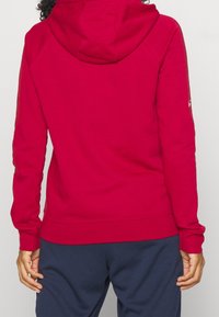 Červená mikina s kapucí z měkké tkaniny, má dlouhé rukávy a žebrovaný lem. Zadní strana ukazuje hladkou texturu a minimalistický design.