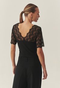 Črna majica z kratkimi rokavi iz zapletene čipke z valovitim vzorcem. Hrbtna stran ima globok izrez v obliki črke V. Ujemajoče se s prilagojenimi črnimi hlačami.