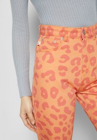 Calças de ganga laranja com estampado de leopardo rosa, apresentando uma cintura alta, acessórios em prata e costuras decorativas. Top de manga longa canelado visível.