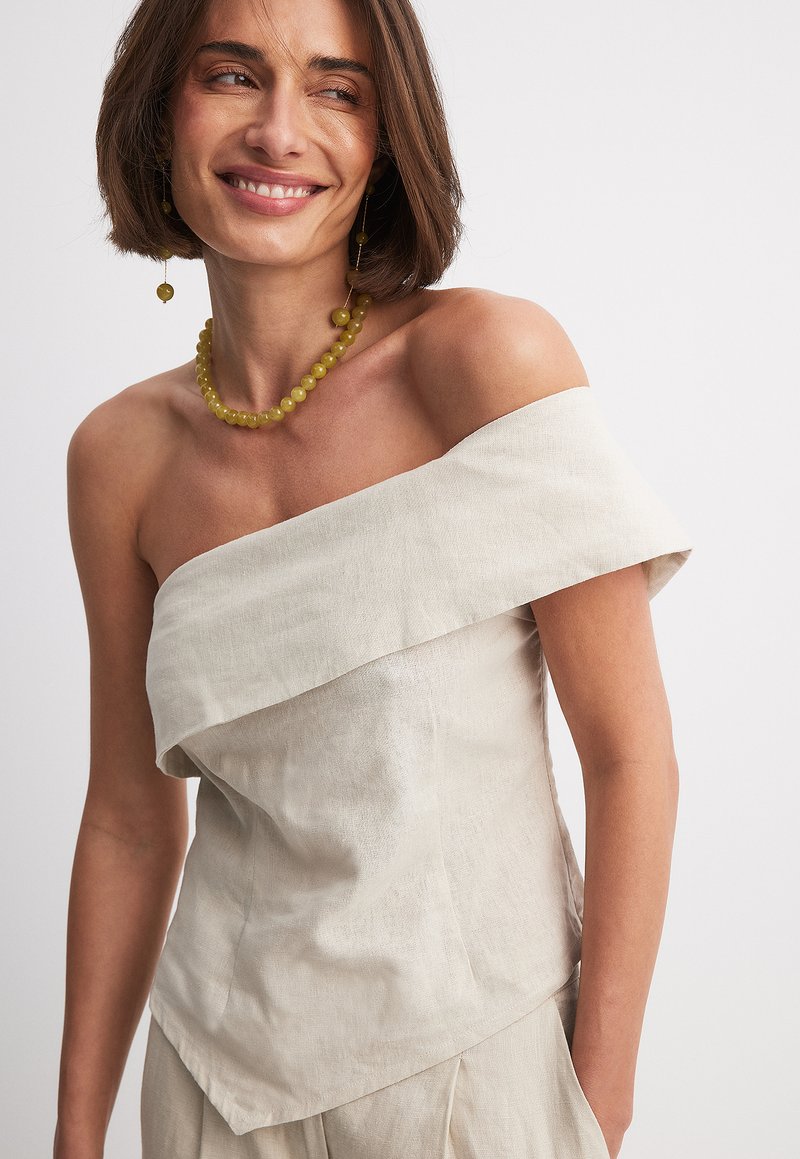 Haut en lin léger beige à épaules dénudées avec un design superposé. Présente une texture lisse et un ourlet asymétrique. Accentué de perles vertes.