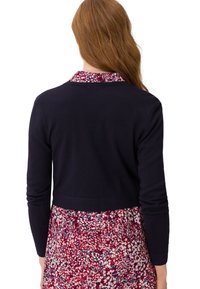 Femme aux longs cheveux roux portant un pull court noir à manches longues par-dessus une robe fleurie rouge, blanche et bleue, vue de dos.