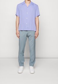 Camicetta button-up a maniche corte, lilla, con colletto rilassato; abbinata a pantaloni azzurri chiari e sneakers bianche, in piedi su uno sfondo semplice.