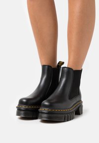 Zwarte leren enkelboots met elastische zijpanelen, chunky rubberen zool, gele stiksels en een trekband aan de achterkant.