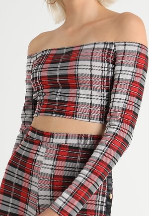 Modèle portant un crop top à carreaux rouges, noirs et blancs, décolleté aux épaules, avec une jupe assortie, sur un fond uni.