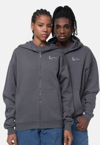 Mørkegrå lynlås hoodie lavet af bomuldsblanding. Har en forkort lomme, ribbede manchetter og kant, samt et broderet logo på brystet.
