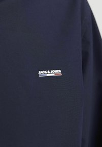 Jūros mėlynos spalvos audinys su lygiu paviršiumi; turi mažą baltą "JACK & JONES" logotipą su raudonais ir mėlynais akcentais. Paprastas dizainas, be raštų.