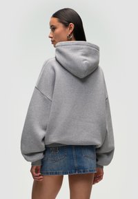 Hoodie grigio oversize con cappuccio, caratterizzato da spalle scese e una consistenza morbida, indossato sopra una mini gonna di jeans blu con una dettaglio del logo.
