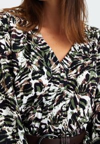 Femme portant un chemisier à col en V avec un imprimé animal abstrait vert, noir et marron, cintré à la taille avec une ceinture cloutée marron foncé.