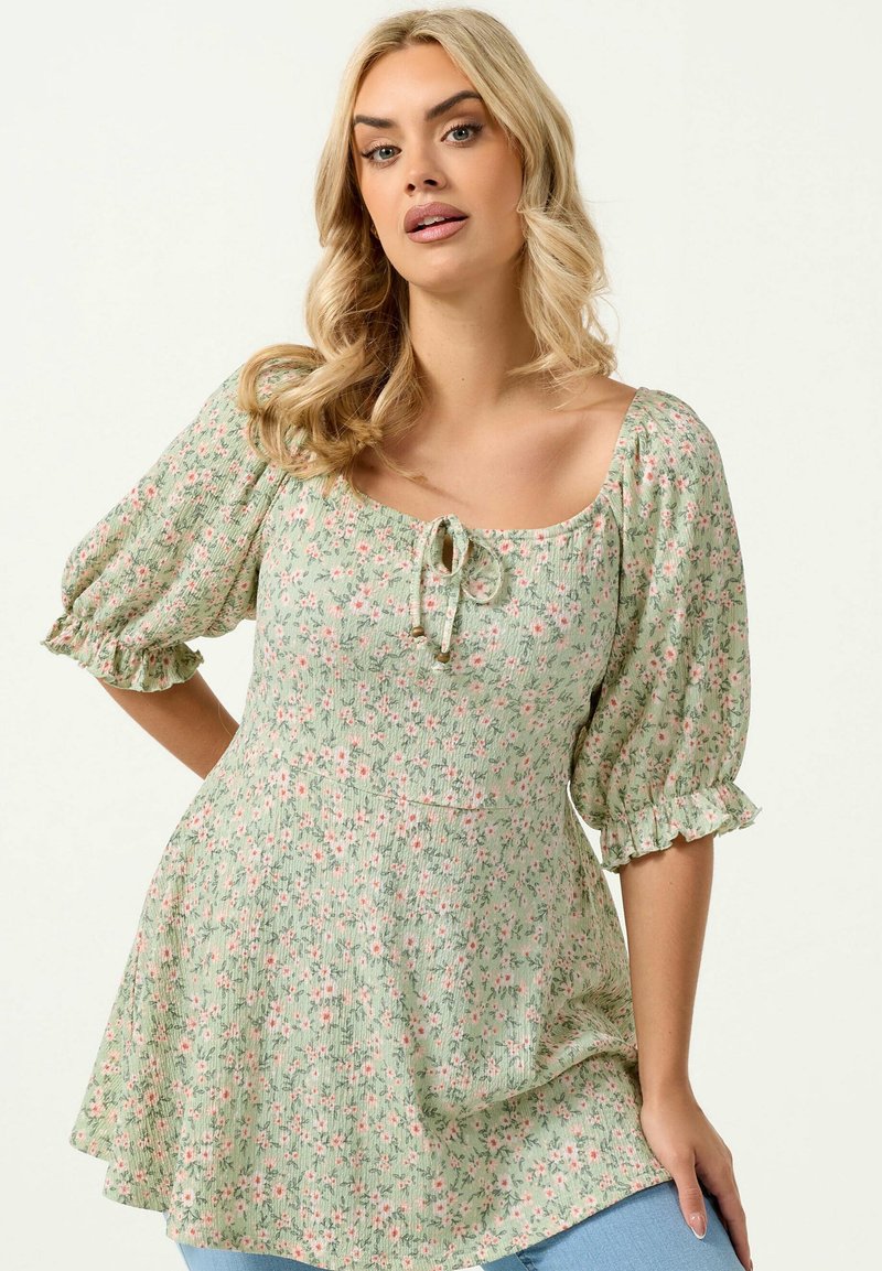 Kobieta o blond włosach w zielonej sukience w kwiaty z bufkami na rękawach i wiązaniem z przodu, pozująca z jedną ręką na biodrze na jednolitym tle.
