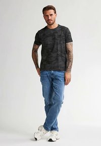 Černé tričko s vzorem a kulatým výstřihem, v kombinaci se světle modrými džínami a bílými teniskami. Model stojí s uvolněnýma rukama.