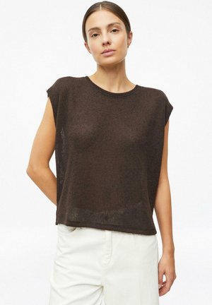 Woman wearing a sleeveless brown knit top and high-waisted white pants, standing with one arm behind her back.