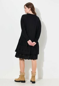 Robe noire à manches longues avec un ourlet en dentelle à œillets superposée. Portée avec des bottes beiges avec une bordure en fourrure. Design simple, texture lisse.