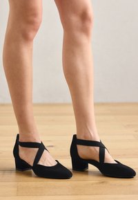 Czarne niskie szpilki z ostro zakończonym noskiem, wykonane z zamszu. Posiadają krzyżujące się paski i otwory na kostki, umieszczone na teksturowanej drewnianej podłodze.