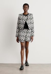 Completo giacca e pantaloni corti a zigzag bianco e nero, realizzato in tessuto testurizzato. Indossato sopra una maglietta nera con scarpe nere. Silhouette semplice.