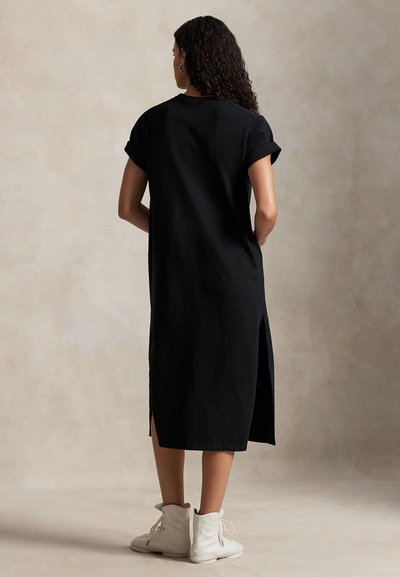 Μαύρο φόρεμα μέχρι το γόνατο με κοντά, γυρισμένα μανίκια, πλαϊνές σχισμές και λεία υφή, συνδυασμένο με λευκά παπούτσια υψηλού πάνω μέρους.