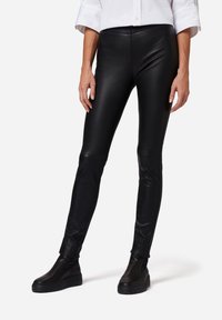 Zwarte nep leren leggings met een slim fit, voorzien van een hoge tailleband en een gladde, glanzende textuur. Gecombineerd met zwarte instap schoenen.