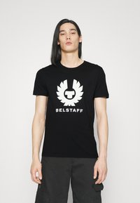 Svart bomullströja med en kontrasterande vit grafik av en stiliserad örn och texten "BELSTAFF" nedanför. Korta ärmar och rund halsringning.