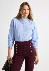 Lichtblauw overhemdsblouse met een gerimpelde kraag en verzamelde mouwen, gecombineerd met bordeauxrode high-waisted broek met gouden knoopaccenten.