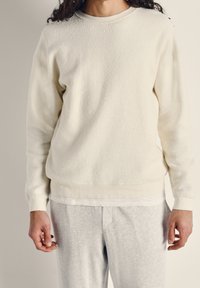 Vit fleece-tröja med en texturerad yta, rund hals och ribbade ärmslut. Buren över ett ljusgrått baslager, ihop med gråa sweatpants.