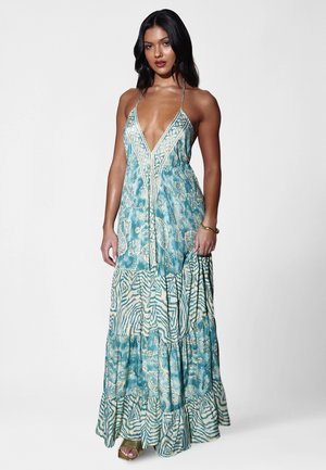 Femme portant une robe longue sans manches bleu sarcelle avec des motifs floraux et géométriques mélangés, un col en V profond et de fines bretelles.