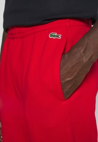 Rote Sweatpants mit elastischem Bund, Seitentaschen und einem grünen gestickten Logo auf der linken Seite. Glatte Stoffstruktur.