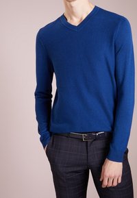 Modrý svetr s výstřihem do V a texturovanou tkaninou, doplněný tmavě kostkovanými kalhotami a černým páskem. Dlouhé rukávy, přiléhavý střih, hladký povrch.