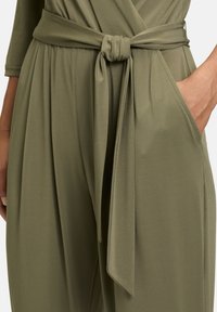 Tuta verde oliva con vita cintata, caratterizzato da una superficie liscia e morbida, maniche lunghe e tasche laterali. Dettaglio a nodo semplice in vita.