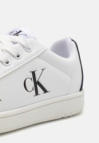 Baskets blanches avec une tige en cuir synthétique lisse, accents noirs sur le talon et un logo "cK" noir proéminent sur le côté. Lacets blancs plats.
