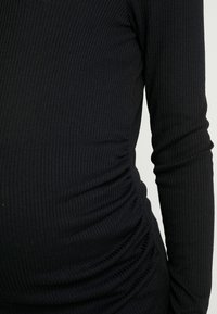 Top preto de manga longa com textura canelada, apresentando um design justo e detalhes franzidos na lateral, realçando uma silhueta curvada.