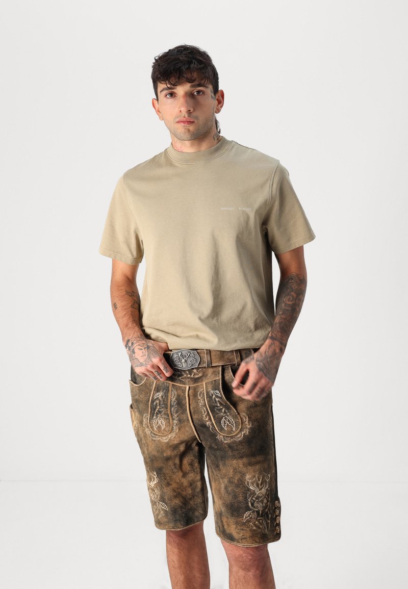 Beige Baumwoll-T-Shirt kombiniert mit braunen, bestickten Shorts mit silbernen Beschlägen und Knopfdetails, getragen mit einer dekorativen Gürtelschnalle.