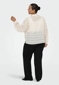 Blusa crema trasparente con motivo geometrico, colletto alto arricciato e maniche ampie, abbinata a pantaloni neri a gamba larga e scarpe nere.