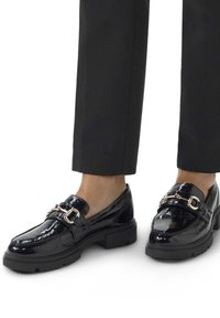 Mocassins en cuir verni noir avec une semelle épaisse et des accents en métal doré. Présente une finition lisse et brillante ainsi qu'une silhouette classique.