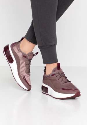 Fødder iført støvede lilla og bordeaux atletiksneakers med hvide såler, parret med mørkegrå joggingbukser, stående på en glat hvid gulv.