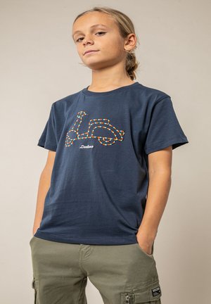 Enfant portant un t-shirt bleu marine avec un motif coloré de scooter Vespa et un pantalon cargo vert olive, mains dans les poches, fond neutre.