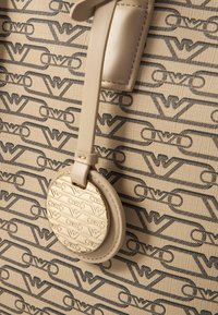 Beige handväska med en strukturerad yta, med ett logomönster i mörka nyanser. Cirkulär metalletikett med präglat logo fäst vid ett band.