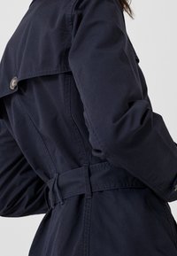 Námořnický trenčkot s páskem v pase, ramenními epauletami a knoflíkovými akcenty. Hladká tkanina s čistým, elegantním designem.